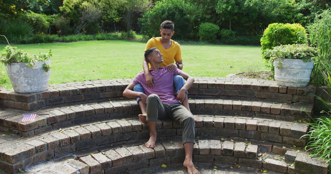 Smiling biracial gay male couple sitting on steps hugging in garden - Free Images, Stock Photos and Pictures on Pikwizard.com