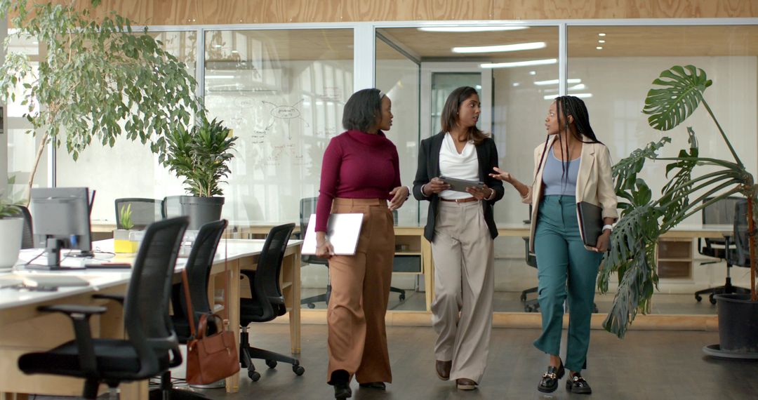 Diverse female coworkers walking and discussing in modern open-plan workspace with plants
