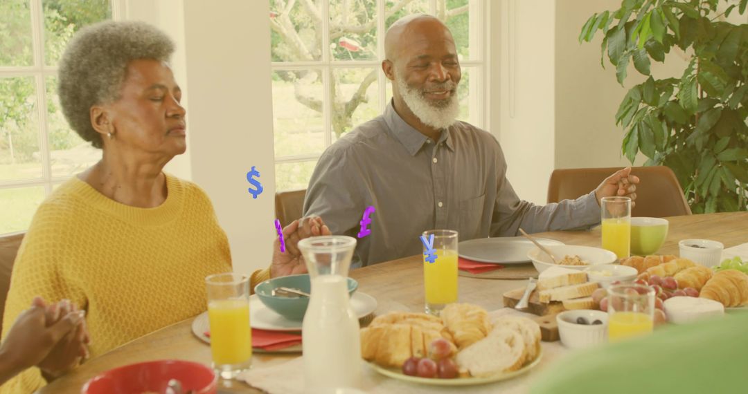 Senior couple holding hands praying at sunlit family breakfast table with croissants and juice