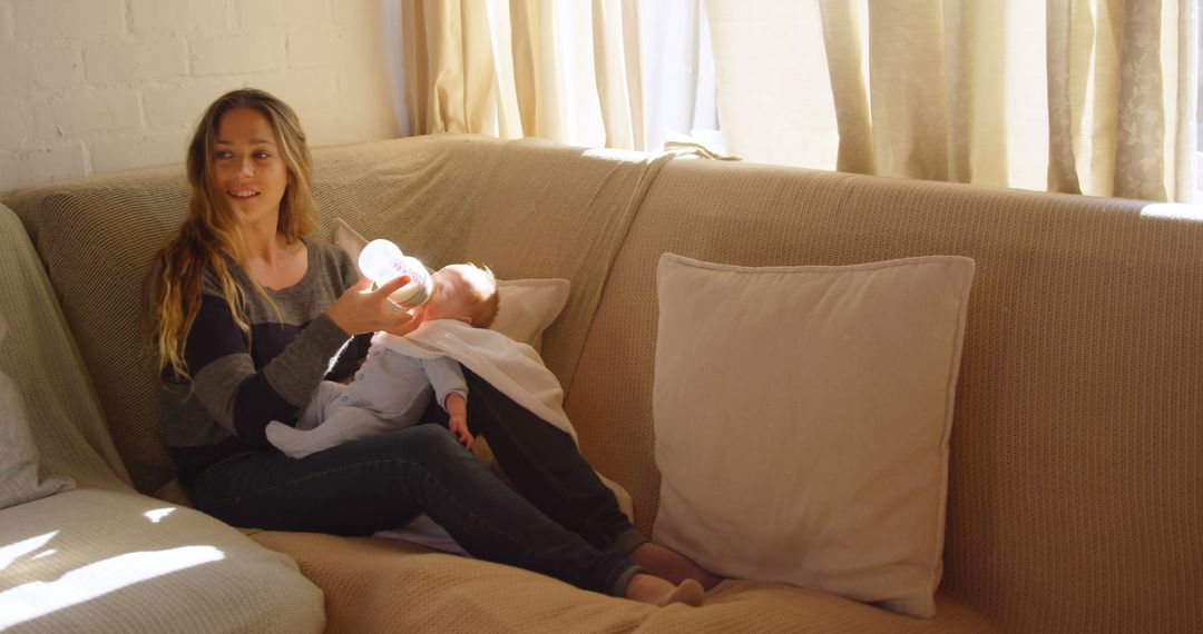 A Caucasian young woman is sitting on a couch, feeding a baby with a bottle, with copy space - Free Images, Stock Photos and Pictures on Pikwizard.com