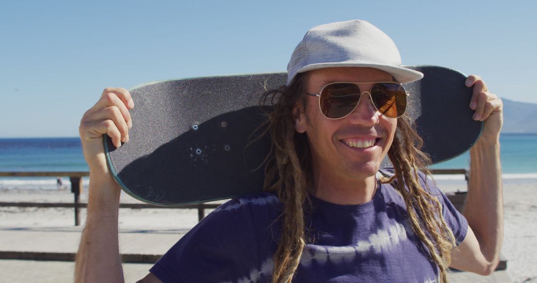 Smiling man with skateboard and dreadlocks at the beach on sunny day - Free Images, Stock Photos and Pictures on Pikwizard.com