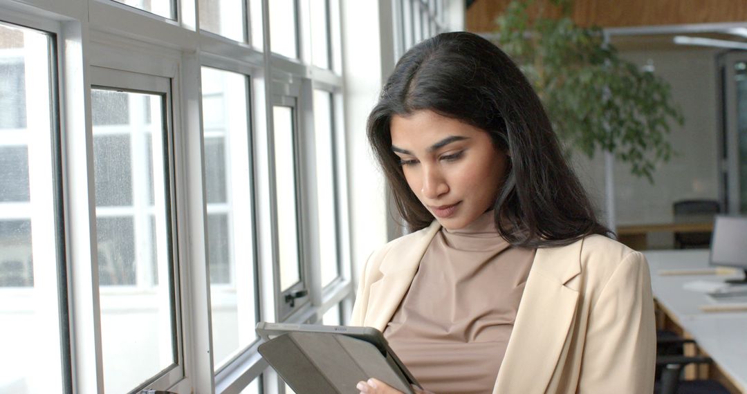 Indian professional using tablet by office windows in beige blazer modern workspace daylight