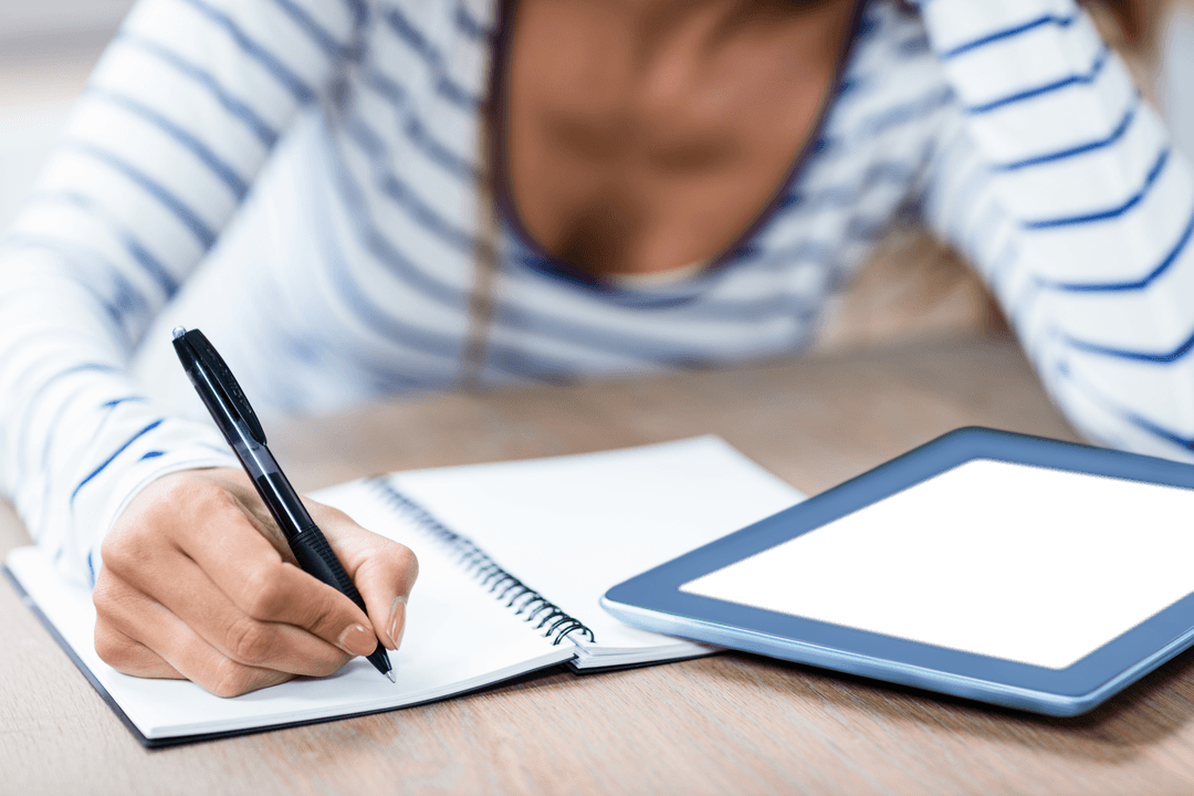 Young Woman Writing on Notebook with Transparent Tablet on Desk
