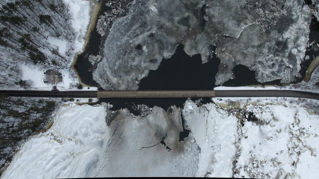 Aerial bridge crossing frozen river with breaking ice and snow-covered forest landscape