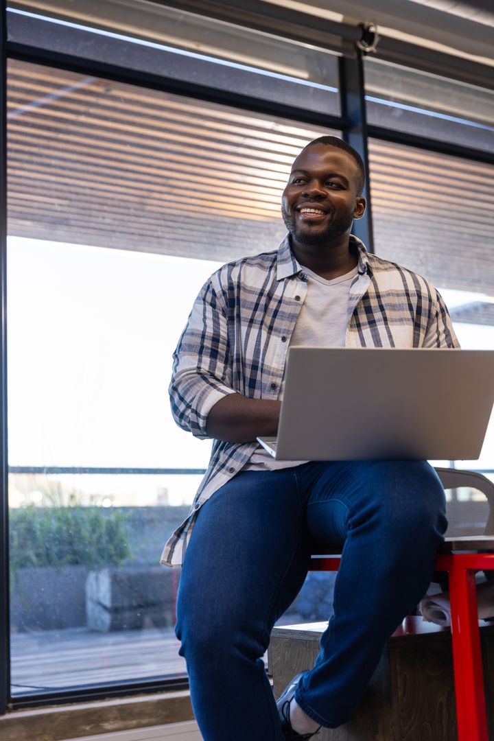 Smiling Man in Casual Workspace Holding Laptop by Window