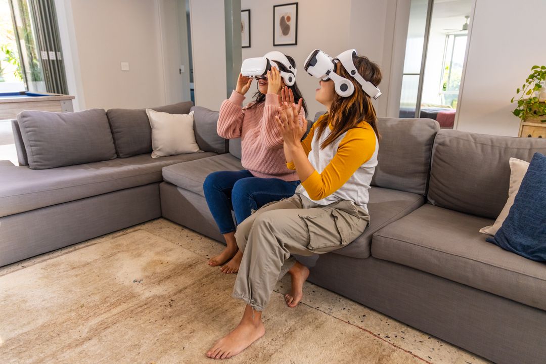 Two Women Using VR Headsets on Cozy Gray Sofa Clapping While Exploring Virtual Reality