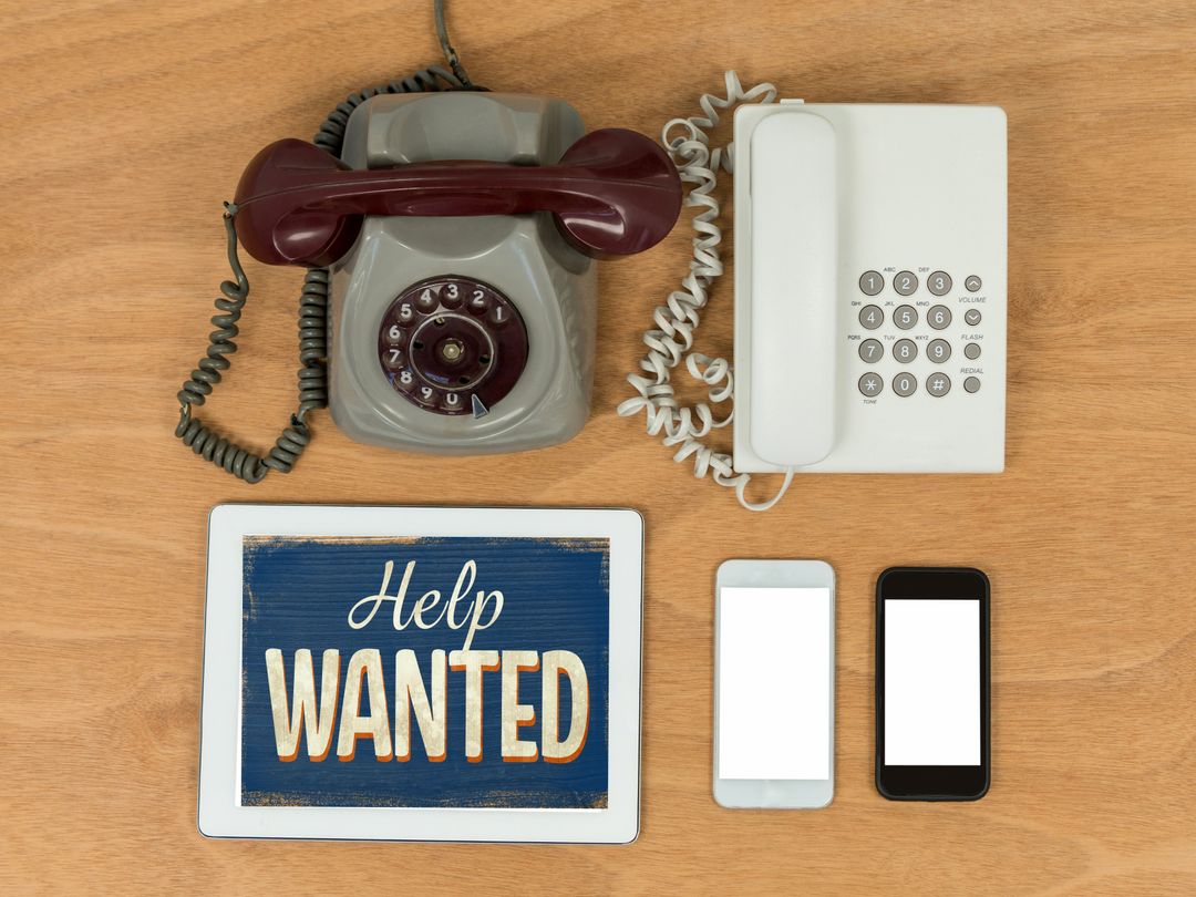 Help Wanted Sign on Tablet Amidst Retro Phones and Smartphones on Wooden Desk - Download Free Stock Templates Pikwizard.com