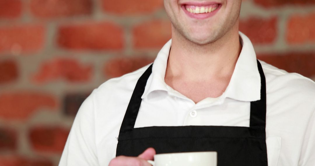 Smiling Barista Holding Mug Wearing Apron Against Brick Wall - Free Images, Stock Photos and Pictures on Pikwizard.com