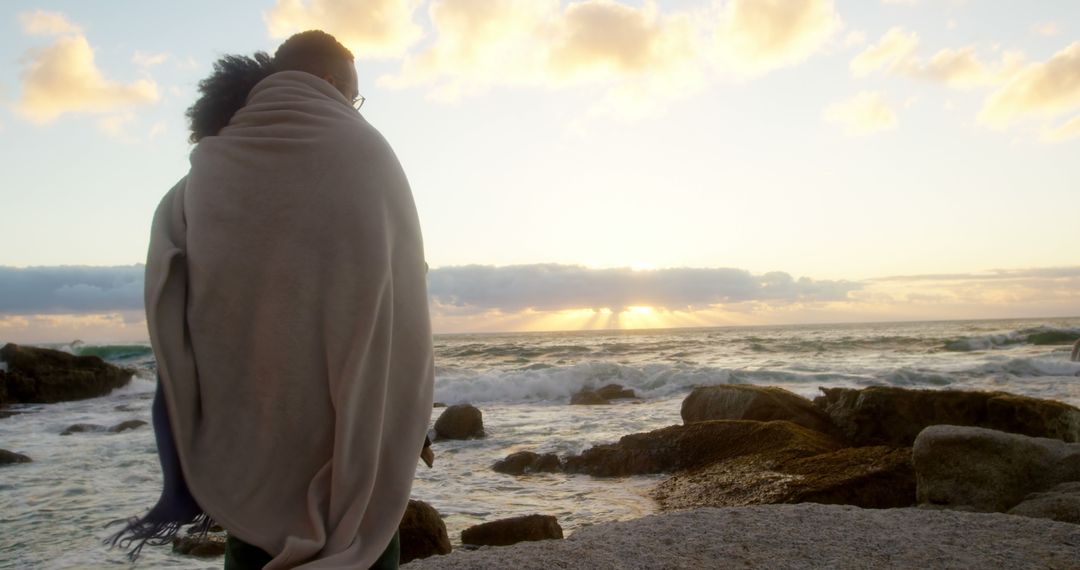 Romantic diverse couple with blankets on backs standing on beach and looking at sunrise - Free Images, Stock Photos and Pictures on Pikwizard.com
