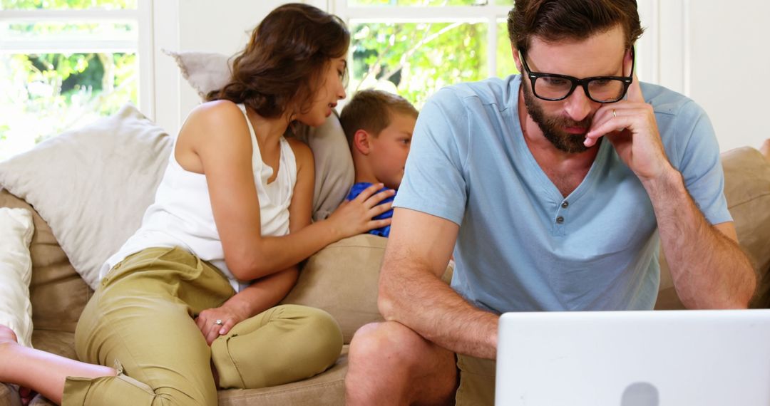 Man Working on Laptop While Family Interacts on Couch in Living Room - Free Images, Stock Photos and Pictures on Pikwizard.com