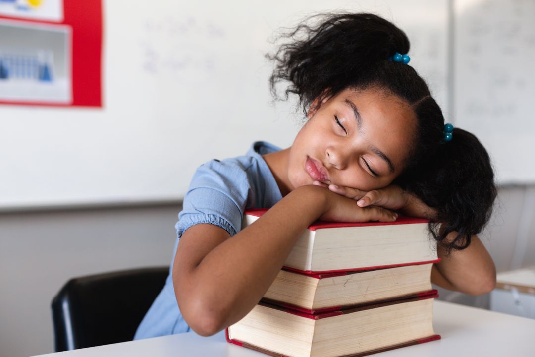 Biracial elementary schoolgirl sleeping on stacked books at desk in classroom - Free Images, Stock Photos and Pictures on Pikwizard.com