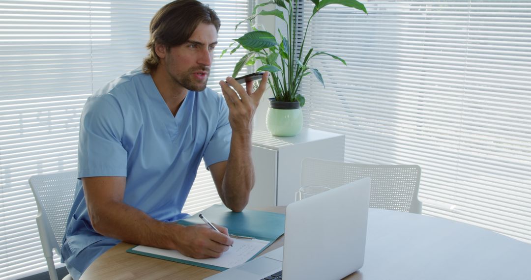 Male doctor in scrubs consulting via phone beside open laptop at office desk - Free Images, Stock Photos and Pictures on Pikwizard.com