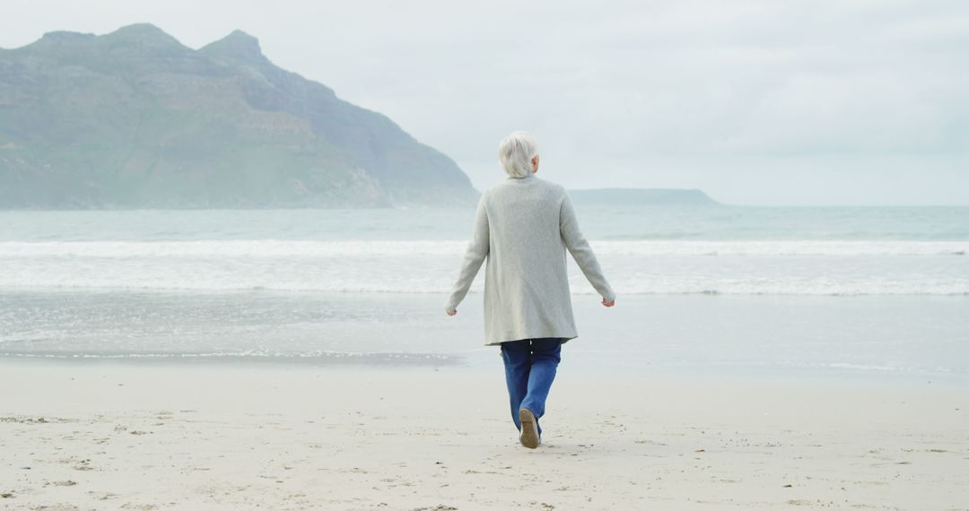 Senior Woman Walking on Tranquil Beach with Mountains in Background - Free Images, Stock Photos and Pictures on Pikwizard.com