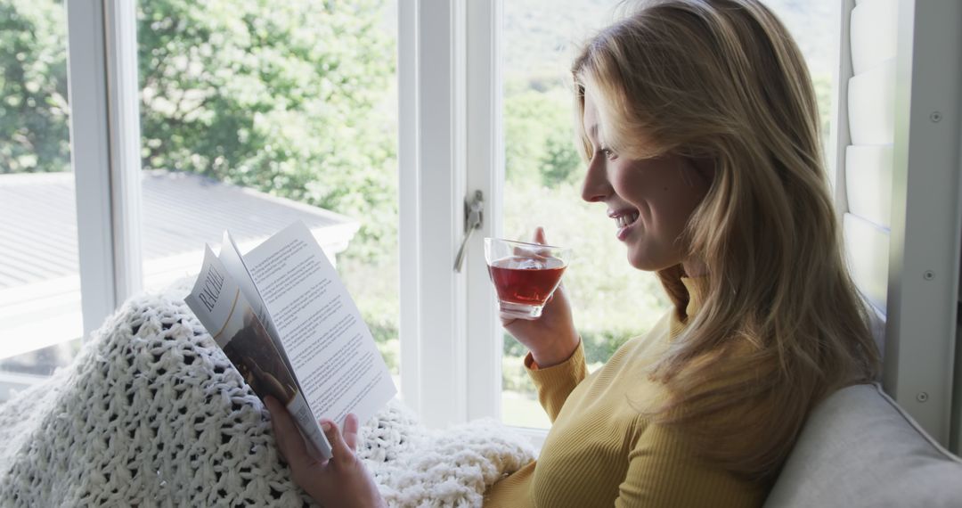 Woman Enjoying Reading Book by Sunny Window with Drink in Hand - Free Images, Stock Photos and Pictures on Pikwizard.com