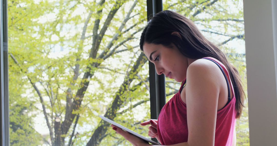 Young Woman Using Tablet Near Window with Green Trees in Background - Free Images, Stock Photos and Pictures on Pikwizard.com