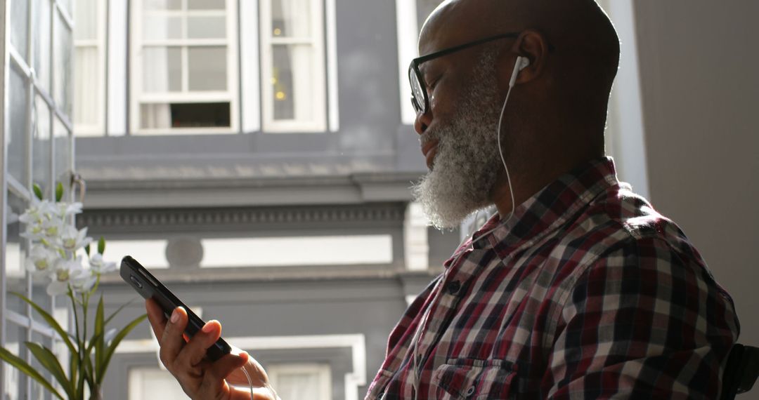 Mature bearded man listening to music on smartphone by window - Free Images, Stock Photos and Pictures on Pikwizard.com