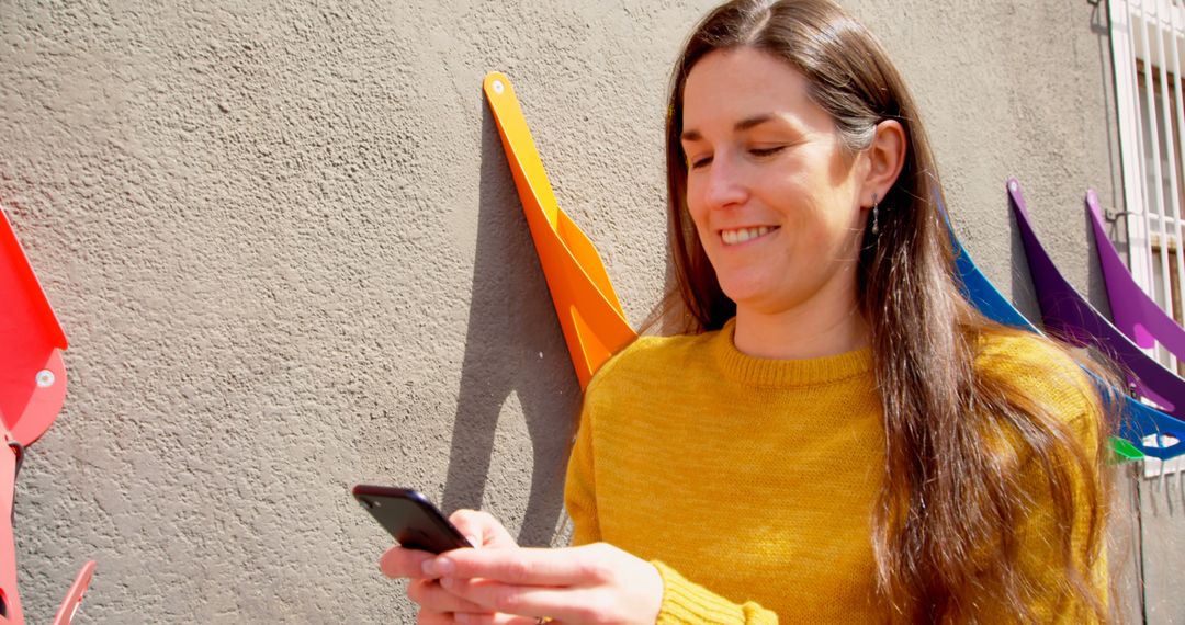 Woman smiling using smartphone next to colorful wall decorations - Free Images, Stock Photos and Pictures on Pikwizard.com