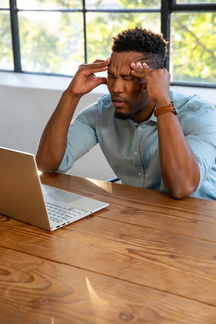 Stressed Man Sitting at Desk with Laptop Rubbing Temples Near Window