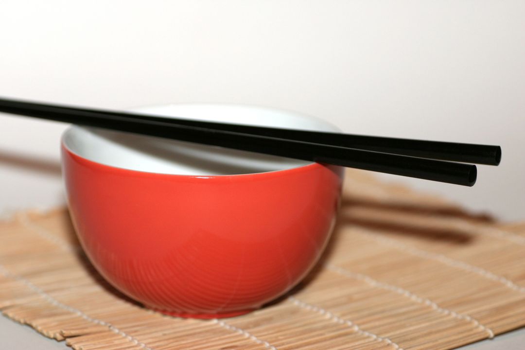 Red Ceramic Bowl with Black Chopsticks Resting on Bamboo Mat for Minimal Tableware