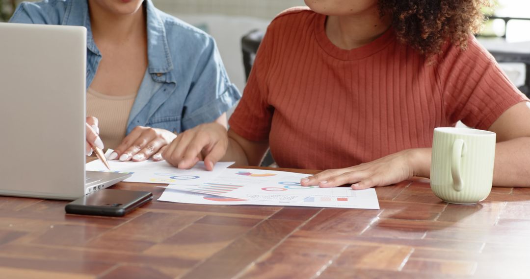 Multiracial women reviewing charts and collaborating on laptop for data analysis at home