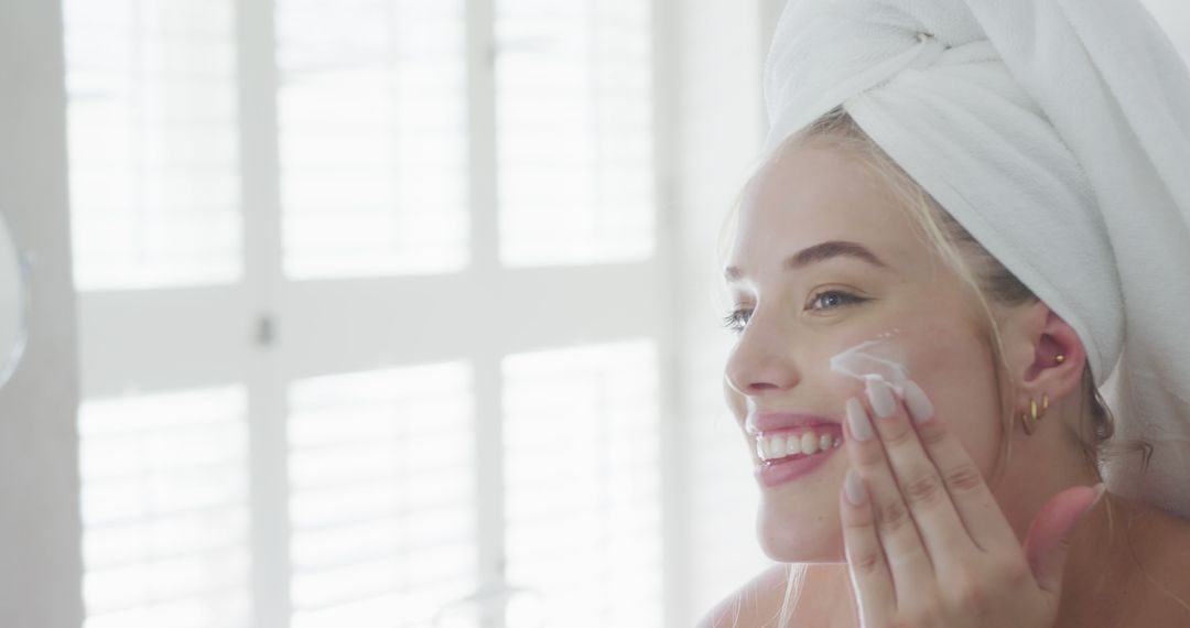 Young Woman Applying Face Cream with Towel Wrapped Hair Smiling in Bright Bathroom - Free Images, Stock Photos and Pictures on Pikwizard.com