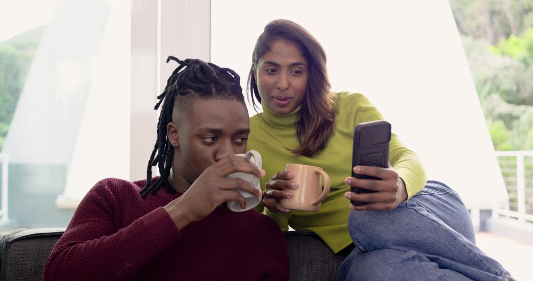 Diverse couple sipping coffee and scrolling phone on sofa sharing cozy morning moment