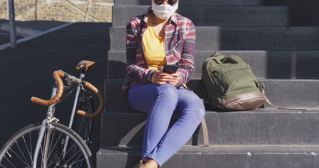 Woman Wearing Mask Sitting on Stairs with Bicycle Beside Her Using Smartphone - Free Images, Stock Photos and Pictures on Pikwizard.com