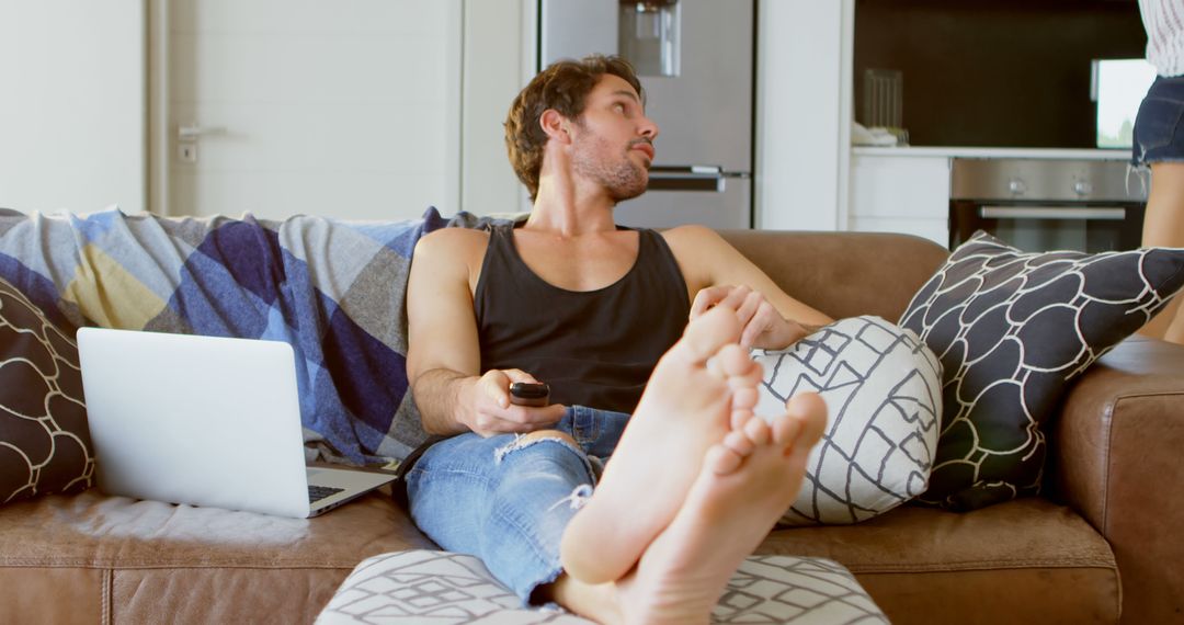 Man Relaxing on Couch with Remote and Laptop, Casual Home Setting - Free Images, Stock Photos and Pictures on Pikwizard.com