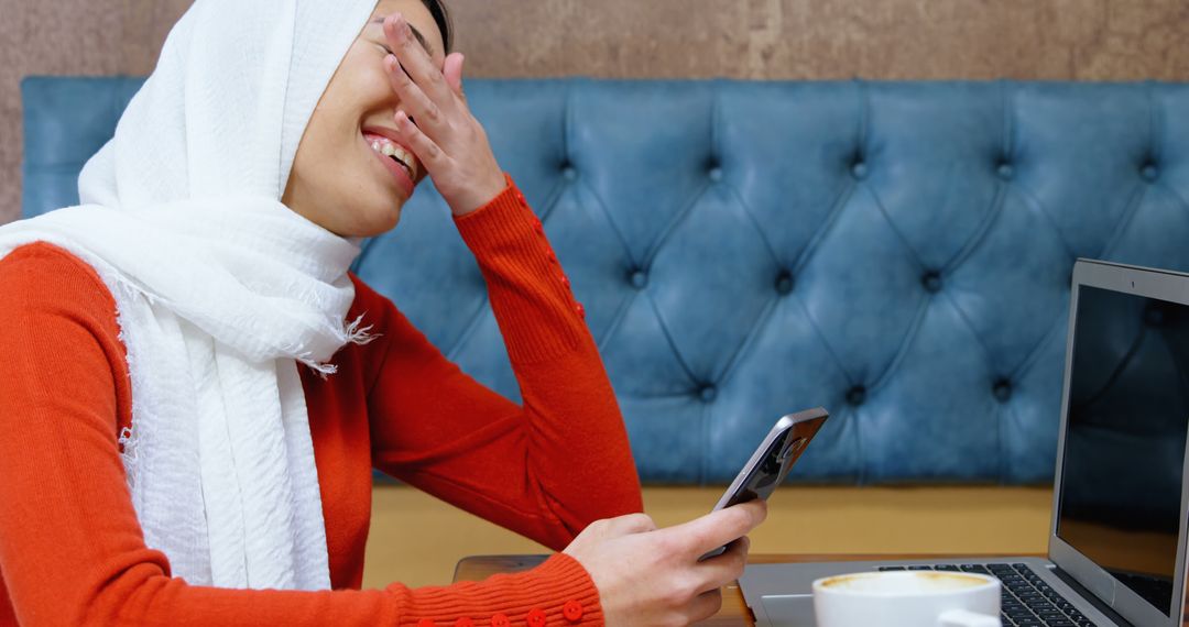 Muslim Woman Laughing While Holding Smartphone Near Laptop in Cafe - Free Images, Stock Photos and Pictures on Pikwizard.com