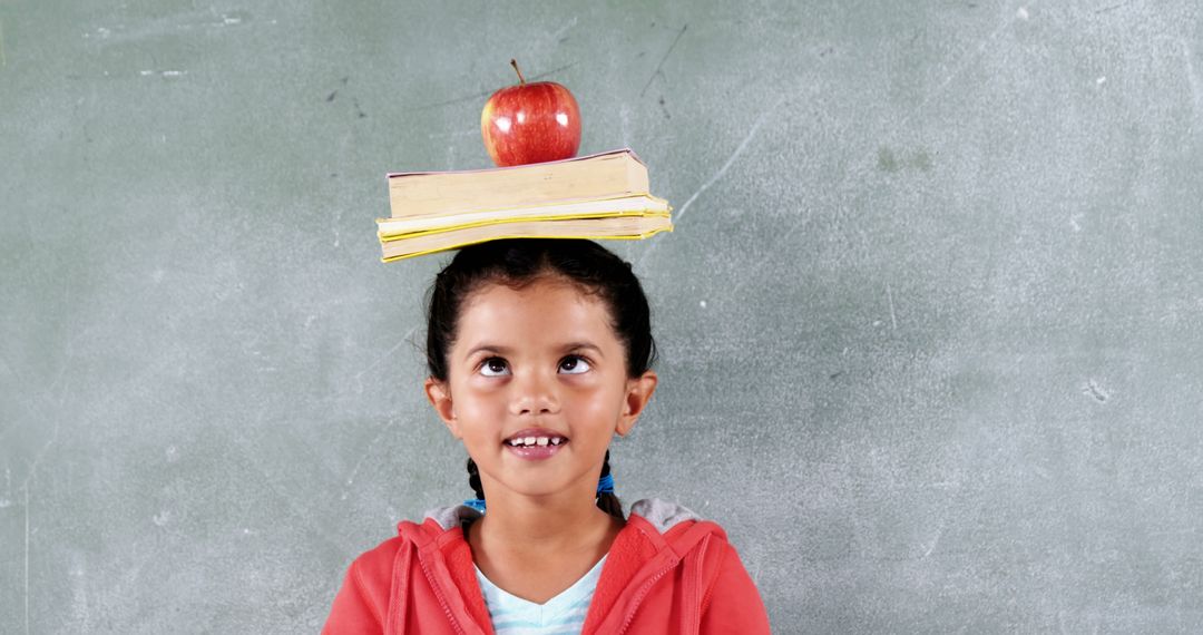 Young Girl with Books and Apple on Head in Front of Chalkboard - Free Images, Stock Photos and Pictures on Pikwizard.com