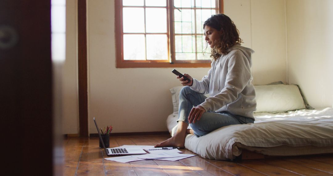 Biracial woman working at home, sitting on bed using smartphone and laptop in cottage - Free Images, Stock Photos and Pictures on Pikwizard.com