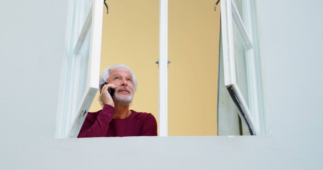 Senior Man Talking on Phone While Looking Out Window in Casual Setting - Free Images, Stock Photos and Pictures on Pikwizard.com