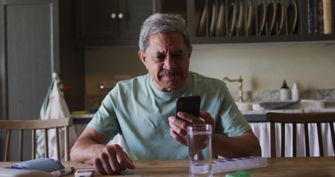 Senior Man Using Smartphone at Kitchen Table with Glass of Water - Free Images, Stock Photos and Pictures on Pikwizard.com