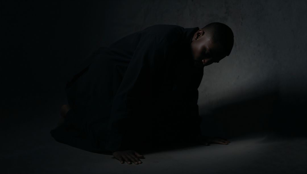 Crouching man in dark robe under dramatic spotlight conveying solitude and introspection