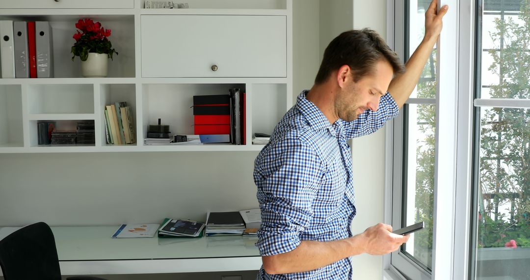 Man checking smartphone while looking out of window in home office - Free Images, Stock Photos and Pictures on Pikwizard.com