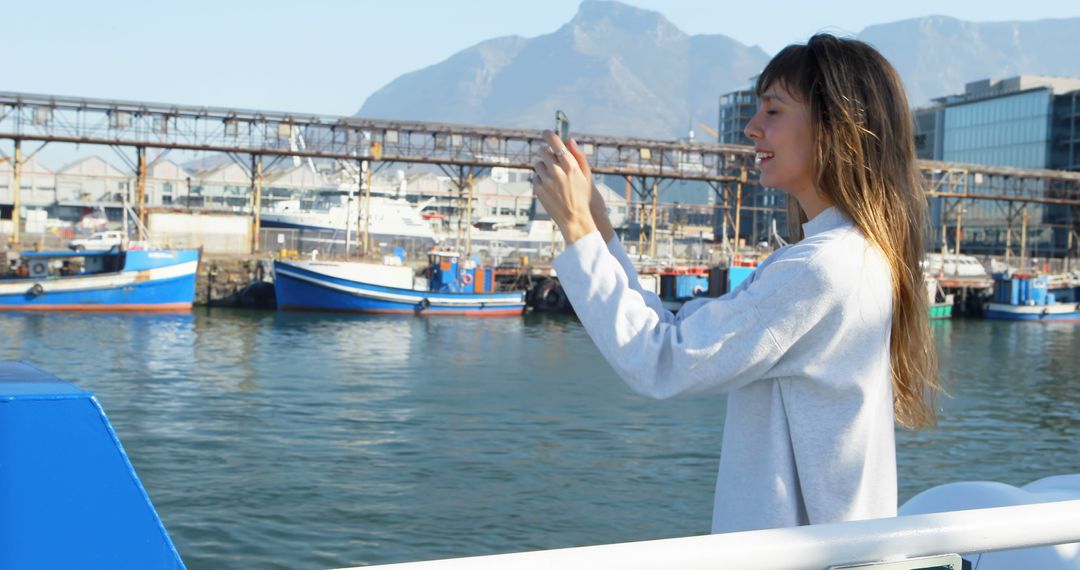 Young Woman Taking Photos at the Harbor with Boats and Mountains in Background - Free Images, Stock Photos and Pictures on Pikwizard.com