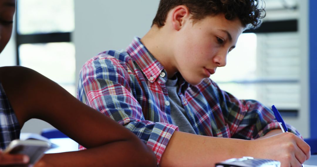 Focused Student Writing with Mobile Phone Nearby in Classroom