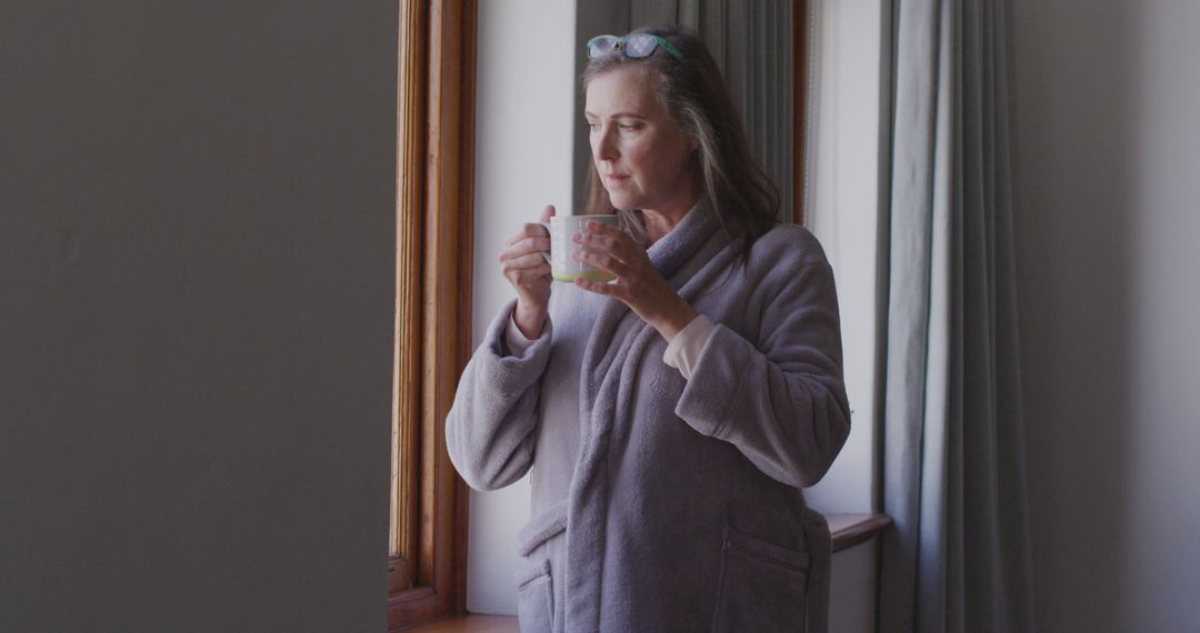 Senior woman in robe enjoying coffee by window at home - Free Images, Stock Photos and Pictures on Pikwizard.com