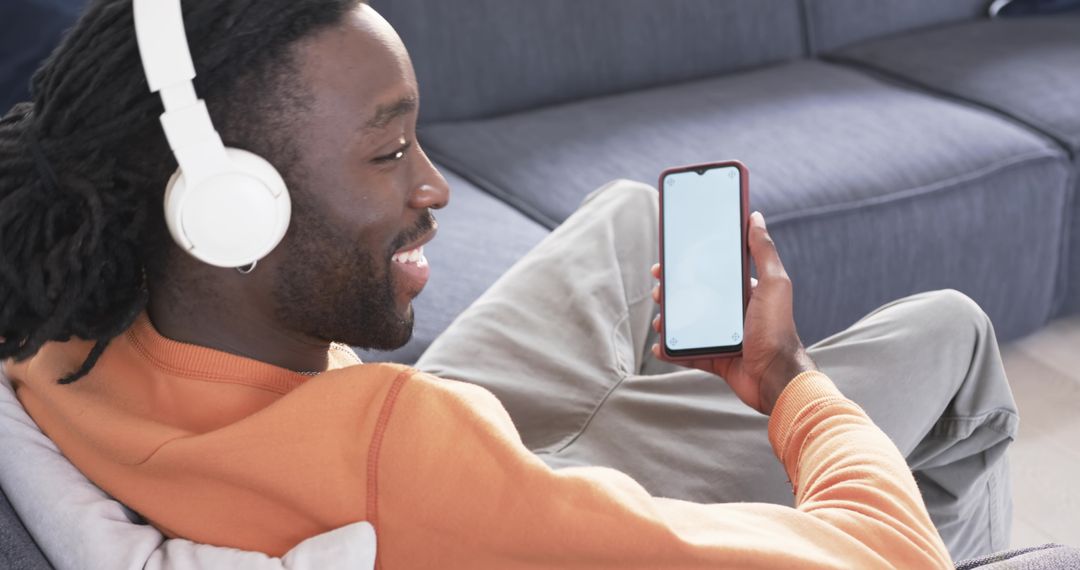 Young man relaxing on sofa wearing white headphones and using smartphone