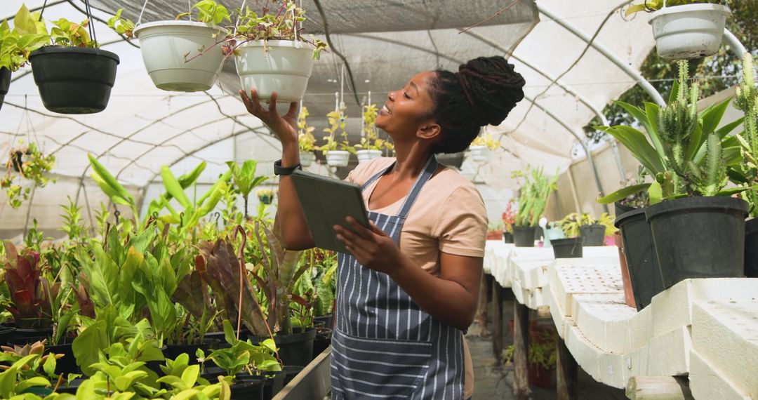 Woman in Apron Inspecting Plants in Greenhouse with Tablet
