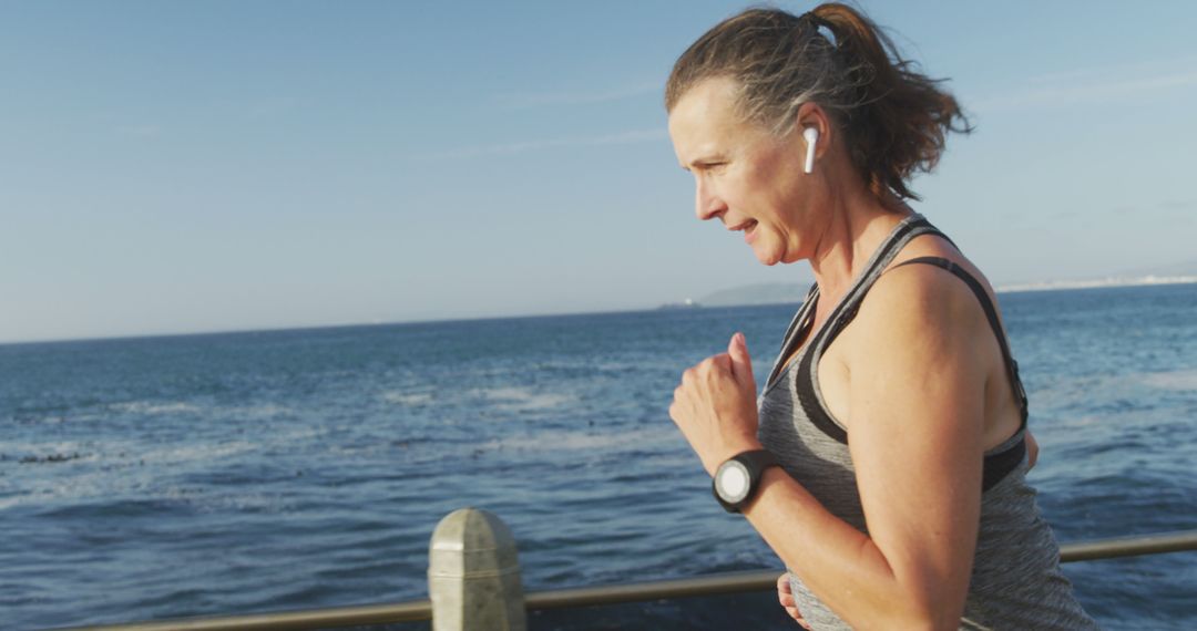 Middle-aged woman jogging by ocean wearing wireless earbuds - Free Images, Stock Photos and Pictures on Pikwizard.com