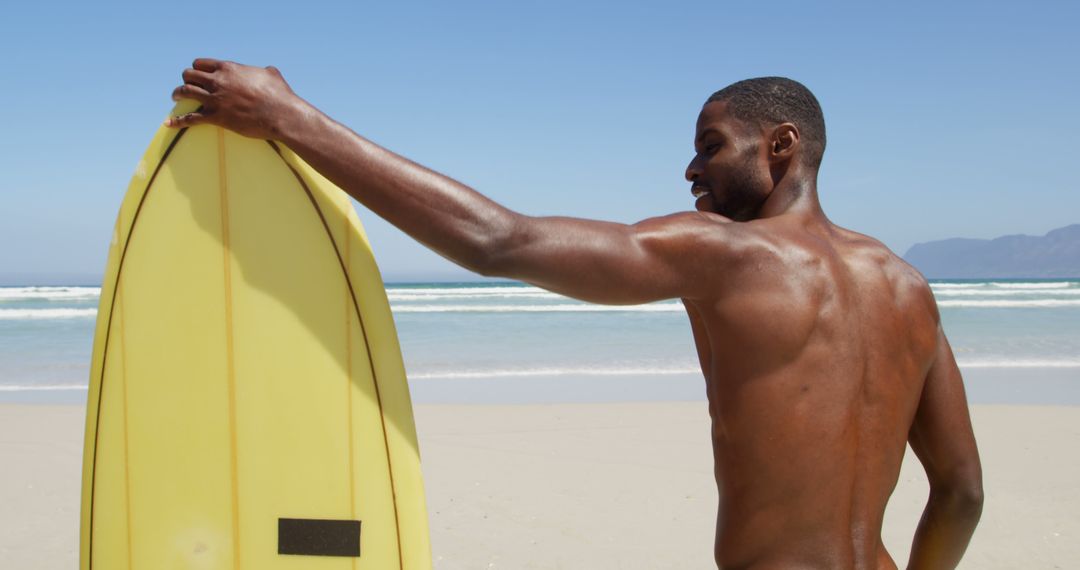 Man Holding Surfboard on a Sunny Beach Looking Towards Ocean - Free Images, Stock Photos and Pictures on Pikwizard.com