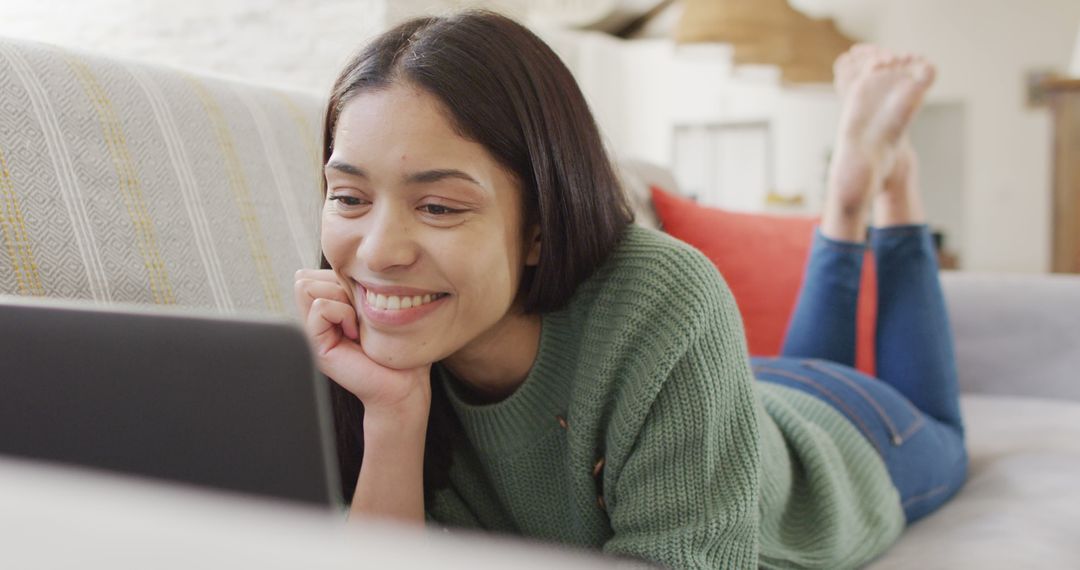 Young woman smiling while lying on a sofa using a laptop at home - Free Images, Stock Photos and Pictures on Pikwizard.com