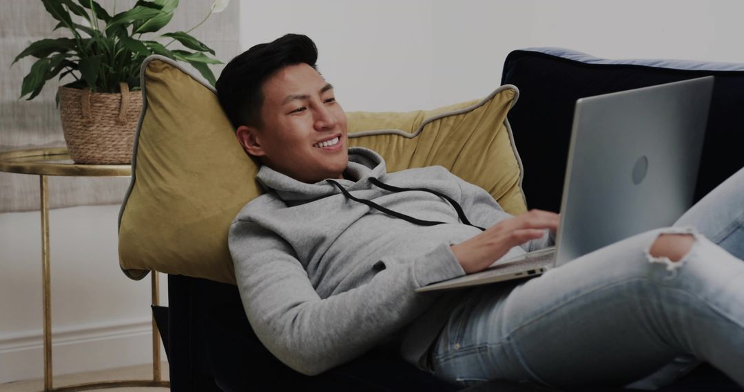 Reclining Asian man typing on laptop on sofa with mustard pillow for remote work and relaxation