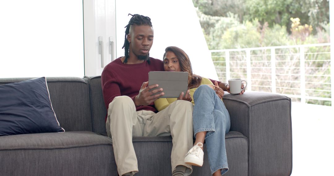 Relaxing diverse couple on sofa using tablet and drinking coffee in bright livingroom