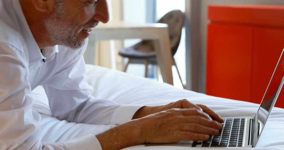 Smiling Mature Man Using Laptop While Lying on Bed in Modern Room - Free Images, Stock Photos and Pictures on Pikwizard.com