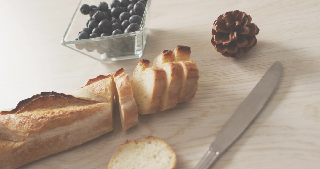 Slicing Baguette with Bowl of Blueberries and Knife on Light Wood Surface