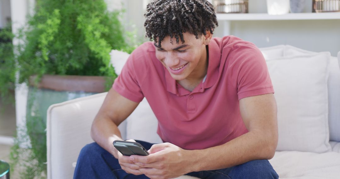 Young man smiling while using smartphone on cozy sofa - Free Images, Stock Photos and Pictures on Pikwizard.com