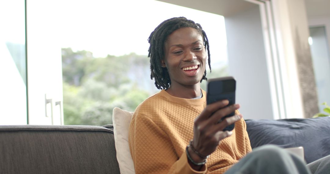 African American man smiling with smartphone on couch by window in mustard sweater