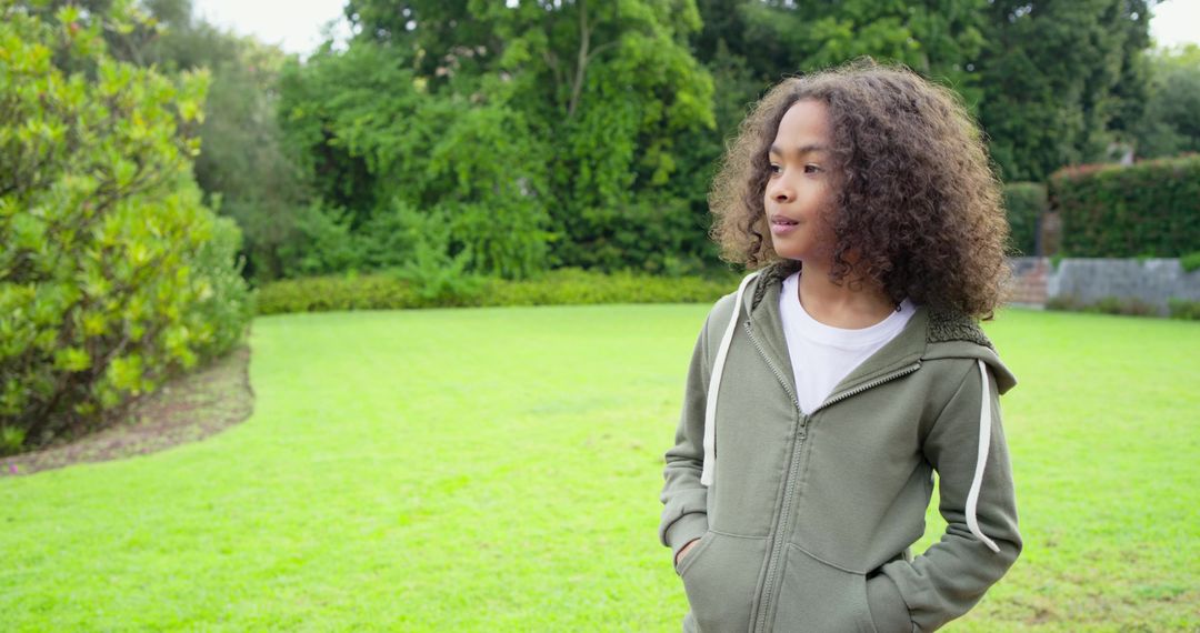 Curly-haired child standing on lawn wearing olive hoodie looking right hands in pockets
